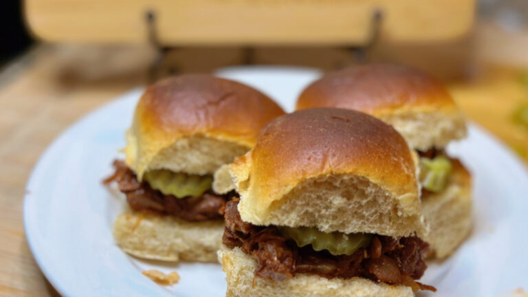 A plate of BBQ jackfruit sliders stacked on soft toasted buns and a drizzle of extra BBQ sauce. The sliders have a rich, smoky color and are served with a side of pickles on a rustic wooden surface.